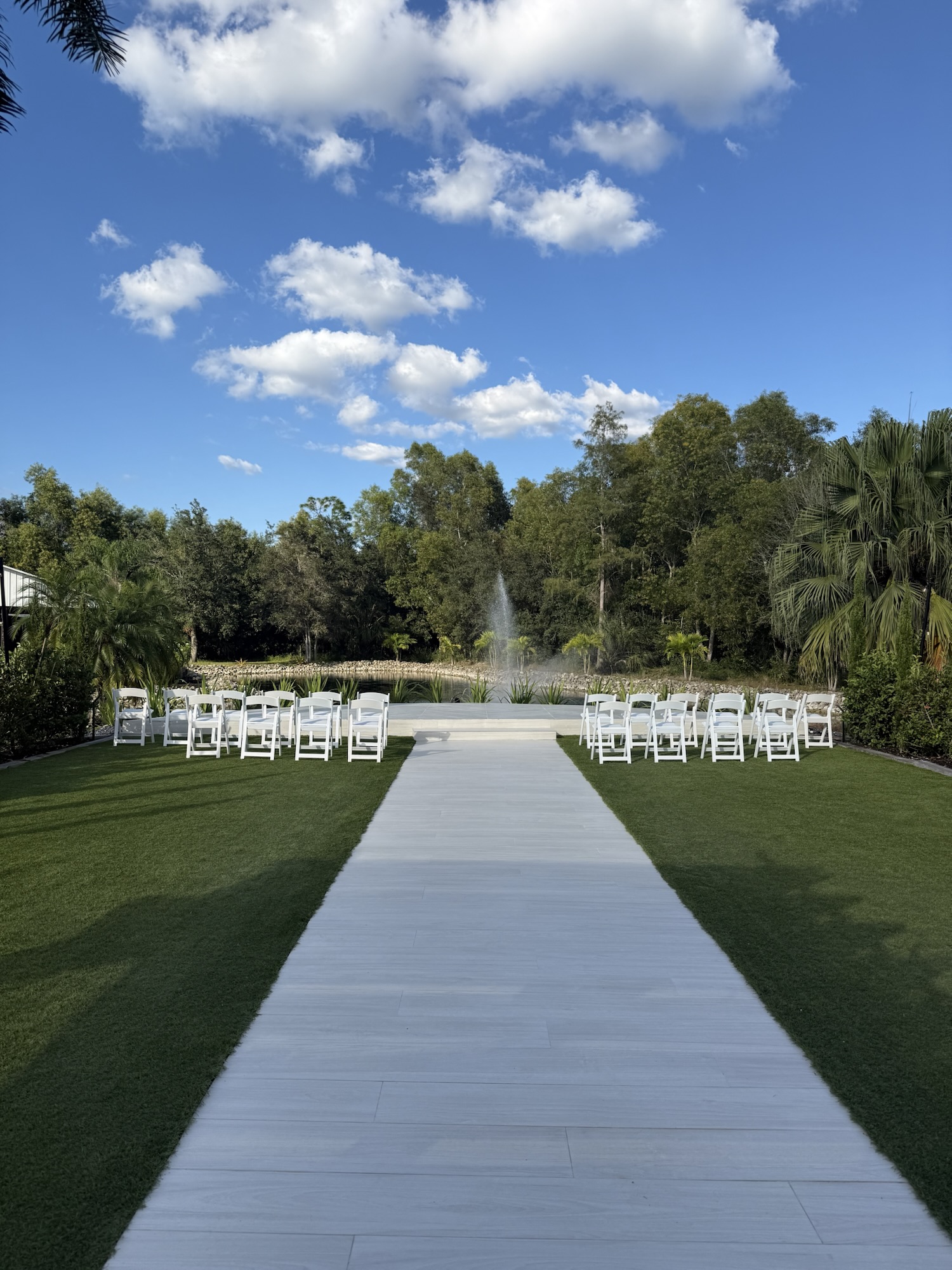 Outdoor wedding ceremony setup with white chairs arranged on either side of an aisle leading to a small stage by a pond with a water fountain and trees in the background under a blue sky with scattered clouds