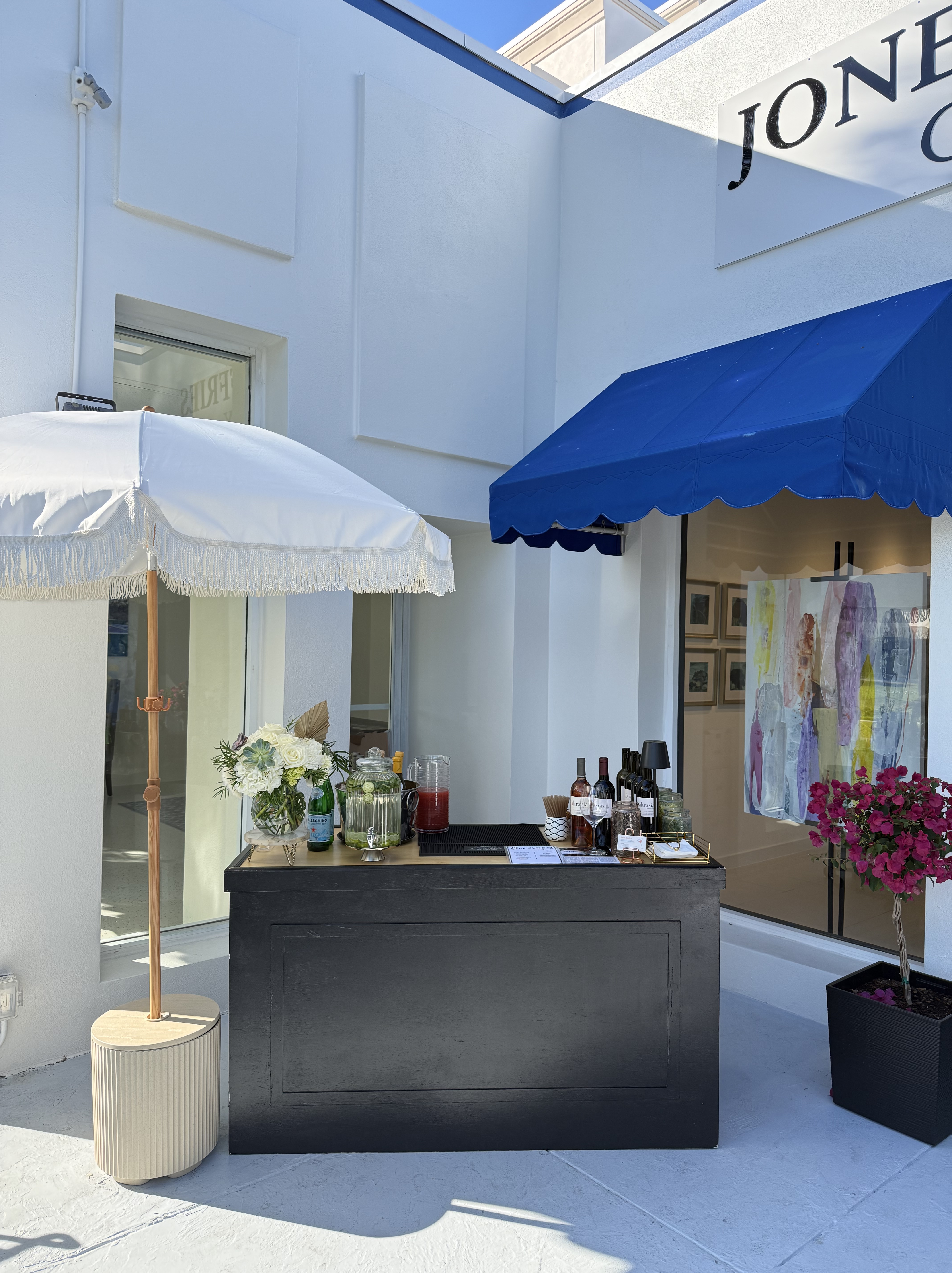 Outdoor beverage station with wine bottles, glass pitchers, flowers, and a white umbrella next to a building with a blue awning and colorful artwork inside
